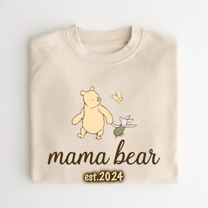 K&ouml;nnte beinhalten: Ein cremefarbener Pullover mit einer Illustration von Winnie Puuh und Ferkel. Der Text "mama bear est. 2024" ist unter der Illustration gedruckt.