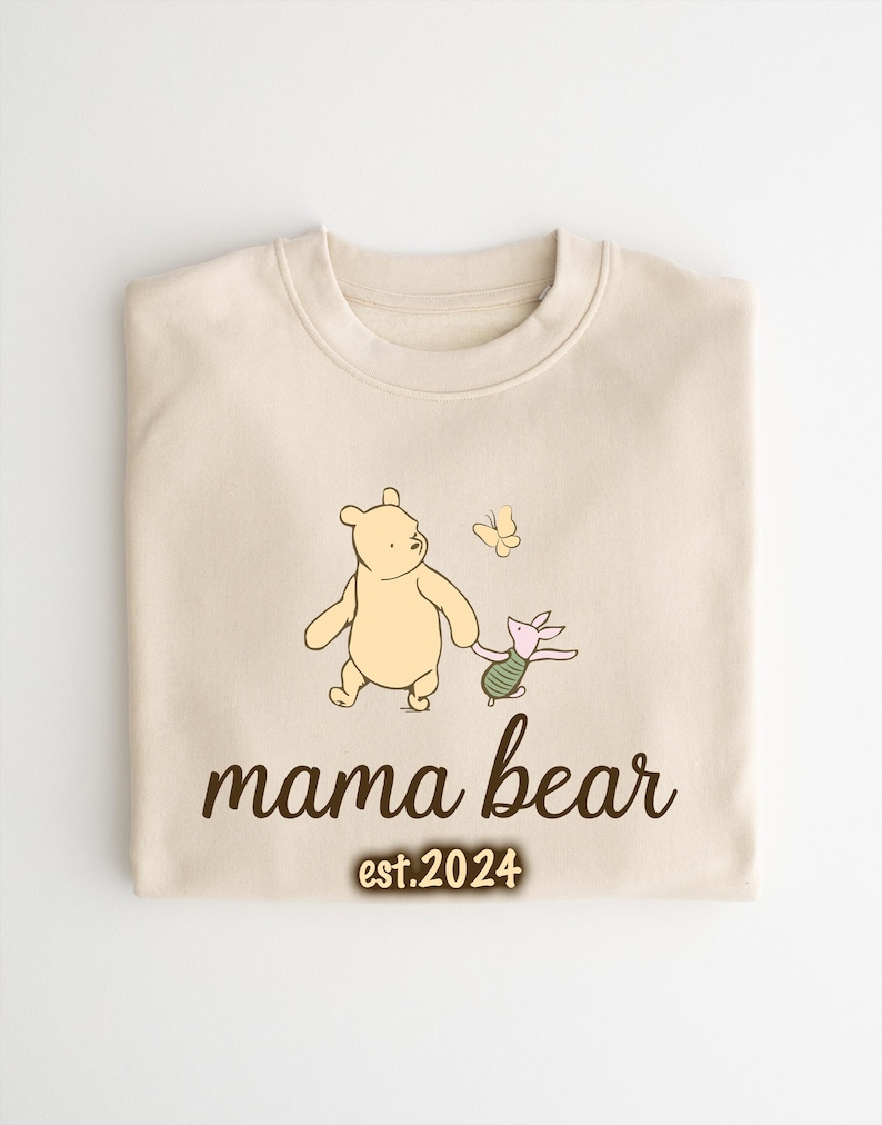 K&ouml;nnte beinhalten: Ein cremefarbener Pullover mit einer Illustration von Winnie Puuh und Ferkel. Der Text "mama bear est. 2024" ist unter der Illustration gedruckt.