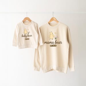 K&ouml;nnte beinhalten: Zwei cremefarbene Sweatshirts mit einem Winnie-the-Pooh-inspirierten Design. Das gr&ouml;&szlig;ere Sweatshirt hat den Text "mama bear est. 2024" und ein Cartoon-Bild von Winnie the Pooh, der mit einem Schmetterling geht. Das kleinere Sweatshirt hat den Text "baby bear est. 2024" und ein Cartoon-Bild von Winnie the Pooh, der mit einem Schmetterling geht.
