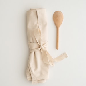May include: A rolled up beige cotton apron with a wooden spoon. The apron is tied with a beige ribbon.