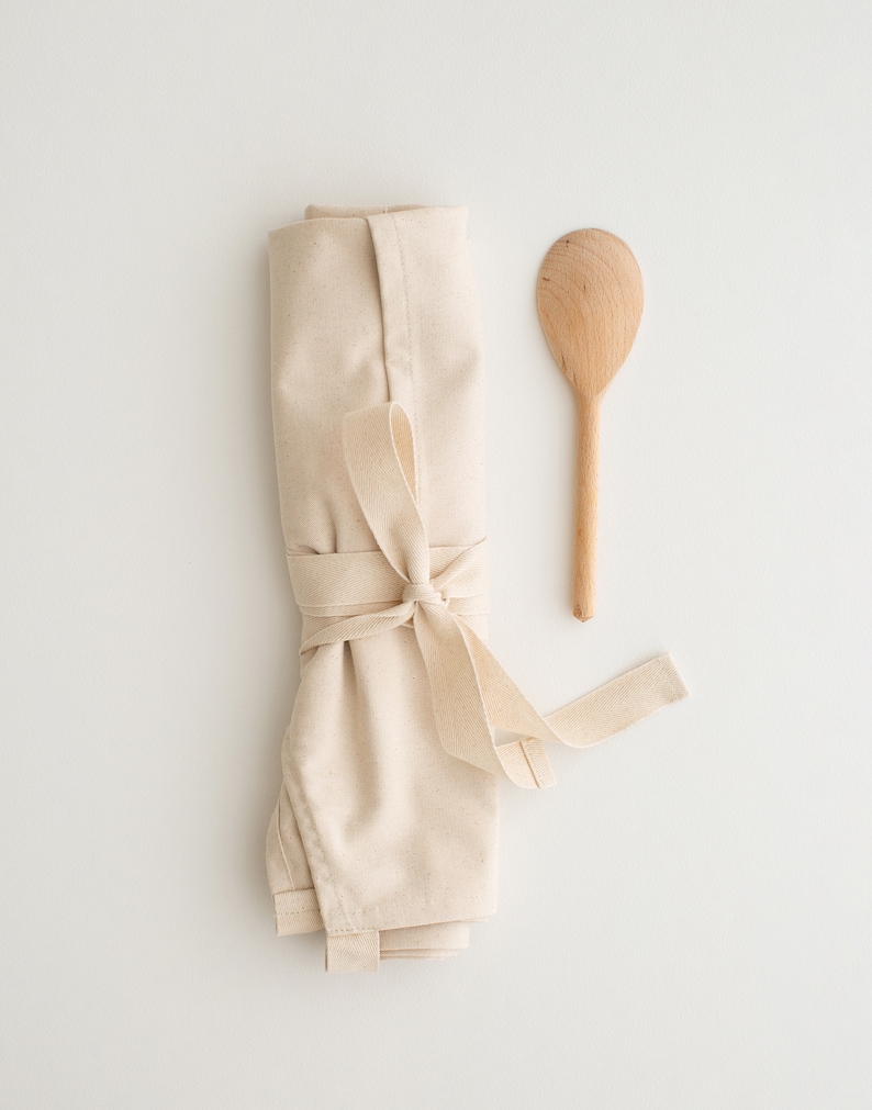 May include: A rolled up beige cotton apron with a wooden spoon. The apron is tied with a beige ribbon.