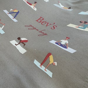 May include: A grey canvas tote bag featuring a repeating pattern of figures in various yoga poses on mats. The center of the bag displays the red text "Bev's" in a bold serif font above the cursive red text "yoga bag" printed on the fabric.