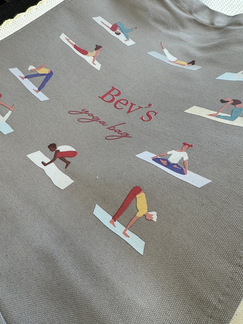 May include: A grey canvas tote bag featuring a repeating pattern of figures in various yoga poses on mats. The center of the bag displays the red text "Bev's" in a bold serif font above the cursive red text "yoga bag" printed on the fabric.