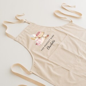 May include: A beige cotton apron with adjustable straps and a large pocket. The apron features a graphic of a cake and other baking supplies with the text "The Perfect Bake Charlotte".