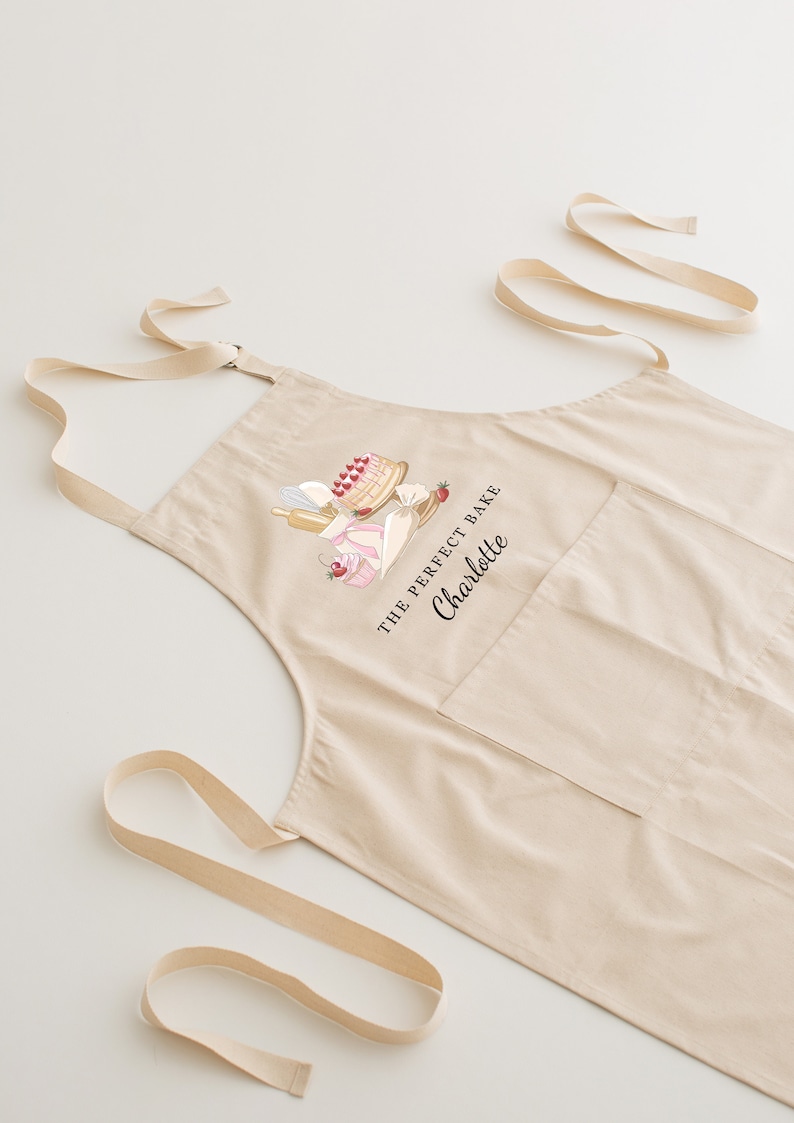 May include: A beige cotton apron with adjustable straps and a large pocket. The apron features a graphic of a cake and other baking supplies with the text "The Perfect Bake Charlotte".
