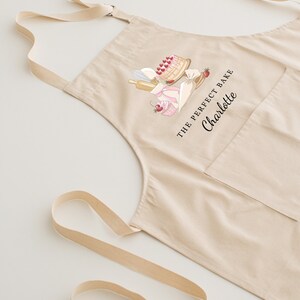 May include: A beige cotton apron with a pocket and adjustable straps. The apron features a graphic of baking supplies and the text "The Perfect Bake Charlotte".