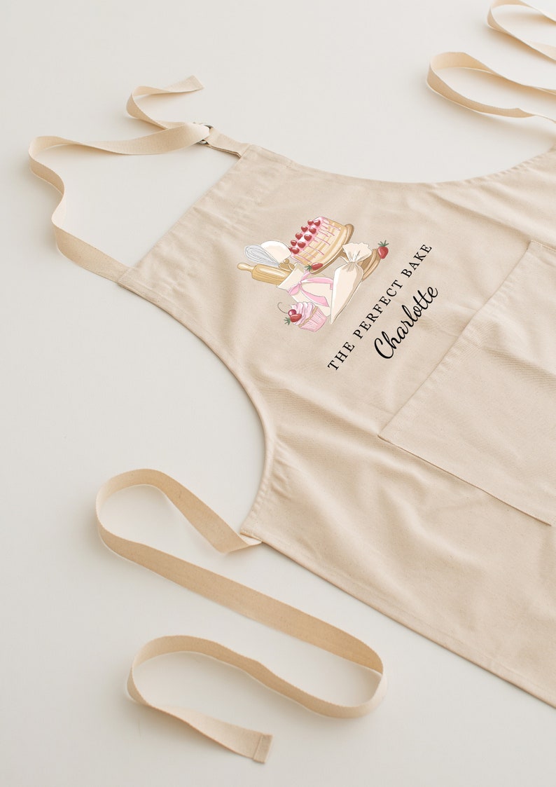 May include: A beige cotton apron with a pocket and adjustable straps. The apron features a graphic of baking supplies and the text "The Perfect Bake Charlotte".