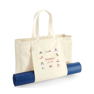 May include: A cream-colored canvas tote bag featuring a graphic print of various yoga poses arranged in a circle around the text "Emma's yoga bag" in red and brown script. A rolled-up dark blue yoga mat is secured horizontally through the bag.