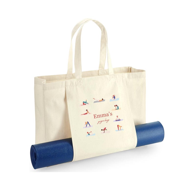 May include: A cream-colored canvas tote bag featuring a graphic print of various yoga poses arranged in a circle around the text "Emma's yoga bag" in red and brown script. A rolled-up dark blue yoga mat is secured horizontally through the bag.