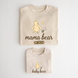 K&ouml;nnte beinhalten: Zwei beige T-Shirts mit Illustrationen von Winnie Puuh und Ferkel. Das gr&ouml;&szlig;ere Shirt tr&auml;gt den Schriftzug "mama bear est. 2024" und das kleinere Shirt "baby bear".