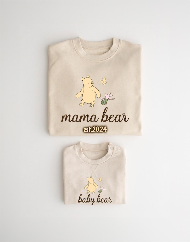 K&ouml;nnte beinhalten: Zwei beige T-Shirts mit Illustrationen von Winnie Puuh und Ferkel. Das gr&ouml;&szlig;ere Shirt tr&auml;gt den Schriftzug "mama bear est. 2024" und das kleinere Shirt "baby bear".