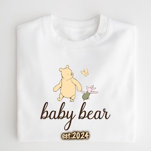 K&ouml;nnte beinhalten: Wei&szlig;er Baby-Strampler mit einer braunen Grafik von Winnie Puuh und Ferkel, die mit einem Schmetterling gehen. Der Text "baby bear est. 2024" ist unter der Grafik gedruckt.