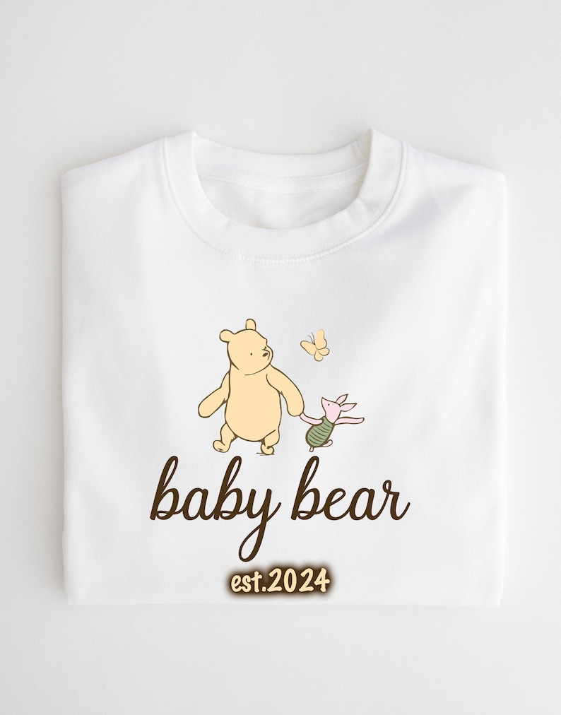 K&ouml;nnte beinhalten: Wei&szlig;er Baby-Strampler mit einer braunen Grafik von Winnie Puuh und Ferkel, die mit einem Schmetterling gehen. Der Text "baby bear est. 2024" ist unter der Grafik gedruckt.