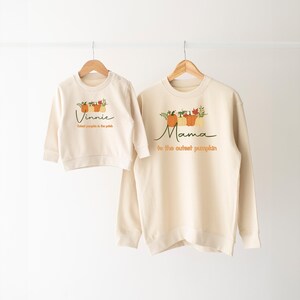 May include: Two cream coloured sweatshirts with pumpkin graphics and text. The larger sweatshirt says "Mama is the cutest pumpkin" and the smaller sweatshirt says "Vinnie cutest pumpkin in the patch".