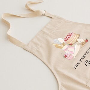May include: A beige cotton apron with adjustable straps. The apron features a graphic of a cake, cupcakes, and baking tools with the text "The Perfect Cake" and "Chef".