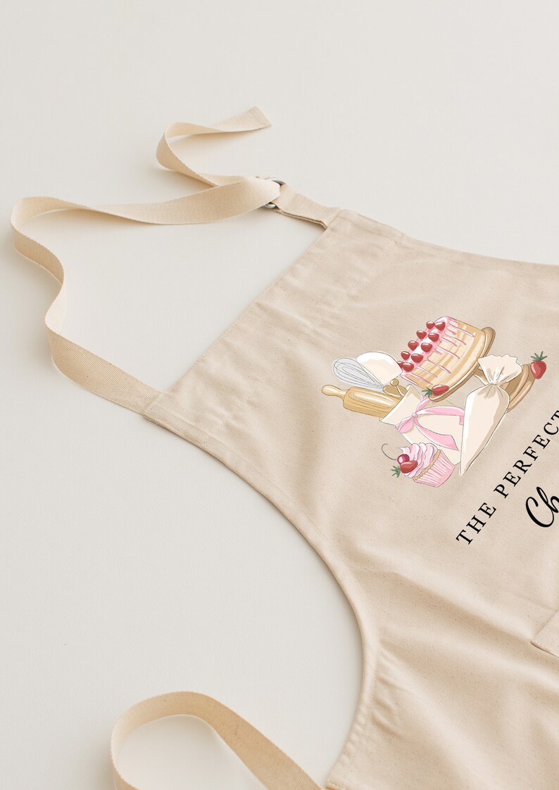 May include: A beige cotton apron with adjustable straps. The apron features a graphic of a cake, cupcakes, and baking tools with the text "The Perfect Cake" and "Chef".