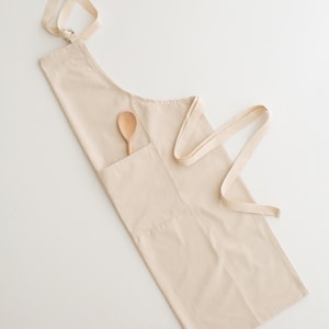 May include: A beige cotton apron with a wooden spoon in the pocket. The apron has adjustable straps and a large pocket.