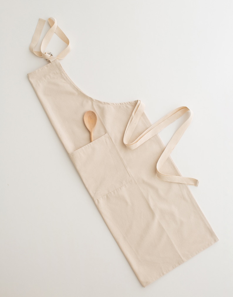 May include: A beige cotton apron with a wooden spoon in the pocket. The apron has adjustable straps and a large pocket.
