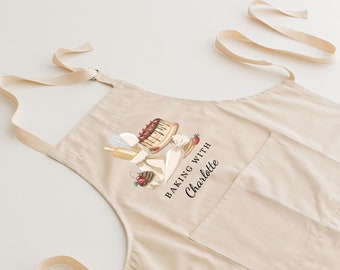 May include: A beige cotton apron with a pocket and adjustable straps. The apron features a graphic of a cake, piping bag, rolling pin, and strawberries. The text "Baking with Charlotte" is printed on the apron.