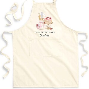 May include: A white cotton apron with a pocket and adjustable straps. The apron features a pink and white illustration of a cake, cupcakes, and baking tools. The text "THE PERFECT BAKE Charlotte" is printed on the apron.