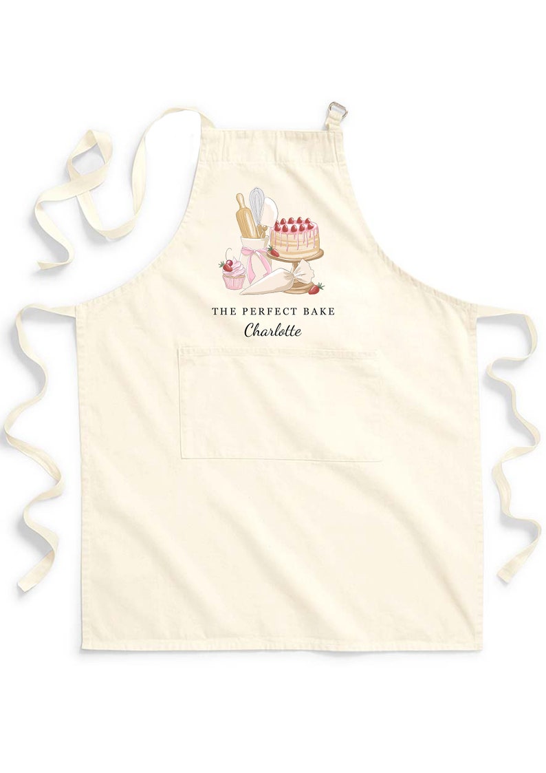 May include: A white cotton apron with a pocket and adjustable straps. The apron features a pink and white illustration of a cake, cupcakes, and baking tools. The text "THE PERFECT BAKE Charlotte" is printed on the apron.