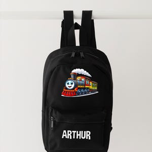 May include: Black backpack featuring a colorful cartoon train with a smiling face and white smoke. The backpack has a front pocket with a zipper and the name "ARTHUR" in white block letters.