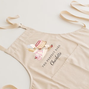 May include: A beige cotton apron with a pocket and adjustable straps. The apron features a graphic of a cake, cupcakes, and a rolling pin with the text "The Perfect Bake Charlotte".