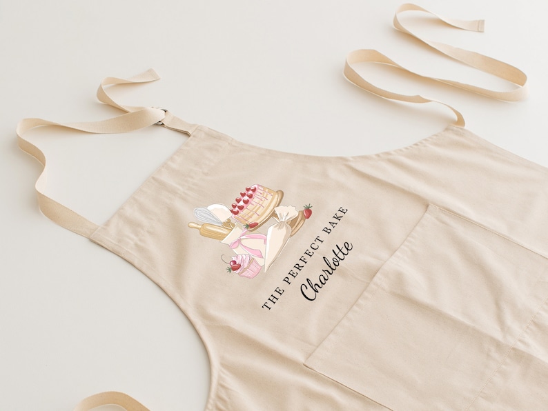 May include: A beige cotton apron with a pocket and adjustable straps. The apron features a graphic of a cake, cupcakes, and a rolling pin with the text "The Perfect Bake Charlotte".