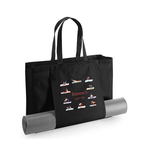 May include: A black tote bag with two handles and a gray yoga mat inserted through the front pocket. The bag features a graphic print of various yoga poses arranged in a circle around the text "Emma's yoga bag" in red and white lettering.