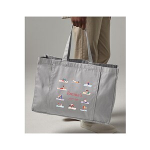 May include: A light gray rectangular canvas tote bag featuring two top handles and a central graphic print. The design displays multiple small figures performing various yoga poses arranged in a circle around the text Emma's yoga bag.