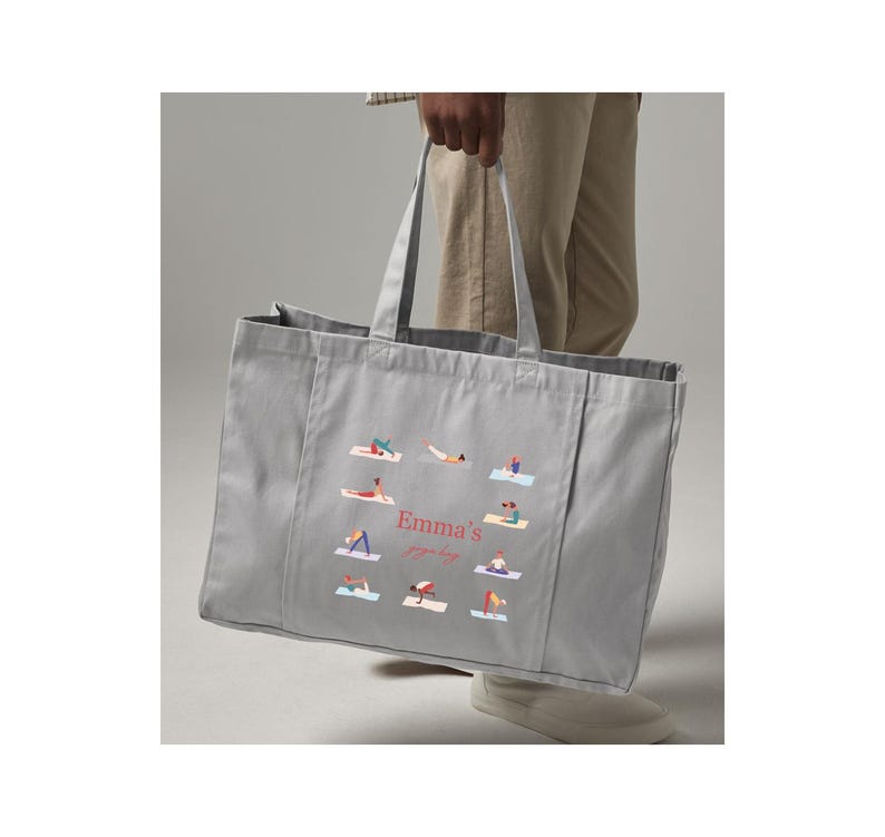 May include: A light gray rectangular canvas tote bag featuring two top handles and a central graphic print. The design displays multiple small figures performing various yoga poses arranged in a circle around the text Emma's yoga bag.