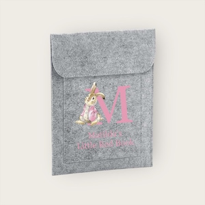 May include: Grey felt book sleeve with a pink letter "M" and a watercolour illustration of a rabbit in a pink jacket. The text "Matilda's Little Red Books" is printed below the rabbit.