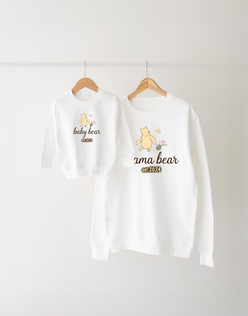 K&ouml;nnte beinhalten: Zwei wei&szlig;e Langarmshirts mit Winnie Puuh-Motiven. Das gr&ouml;&szlig;ere Shirt tr&auml;gt den Schriftzug "mama bear est. 2024" und das kleinere Shirt den Schriftzug "baby bear est. 2024".