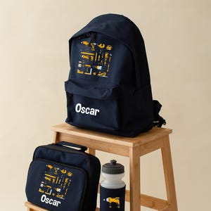 May include: A navy blue backpack, lunch bag, and water bottle set. Each item has a yellow tool graphic and the name "Oscar" in white. The set is displayed on a small wooden stool.