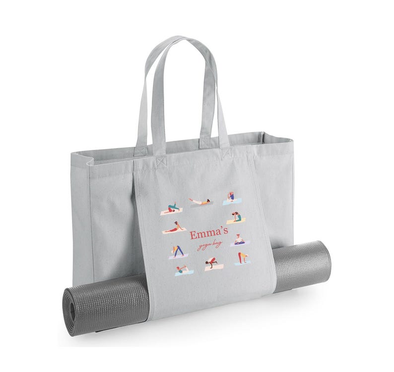 May include: A light gray canvas tote bag with two handles and a horizontal strap across the front designed to hold a rolled-up dark gray yoga mat. The bag features a printed graphic of various yoga poses and the text Emma's yoga bag in red.