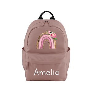 May include: A dusty rose pink backpack with a gold and pink rainbow design and floral accents. The backpack has a white name tag that says "Amelia".