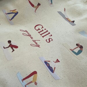 May include: A beige canvas tote bag featuring a repeating pattern of figures in various yoga poses on mats. The center of the bag displays the red text "Gill's yoga bag" in a combination of bold block lettering and a cursive script font.