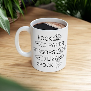 Peut inclure: Mug en céramique blanche avec texte noir et illustrations des gestes de "Pierre, Papier, Ciseaux, Lézard, Spock". Le mug contient un liquide foncé, probablement du café ou du thé. Il est posé sur une surface en bois.