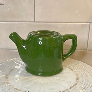 May include: A white cake stand with a gold trim and a green teapot on top. The cake stand has a decorative pattern of small dots and lines.