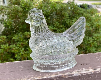 May include: A clear glass hen-shaped dish with a textured surface. The hen is perched on a basket-like base. The dish is sitting on a brown wooden surface, with green foliage in the background.