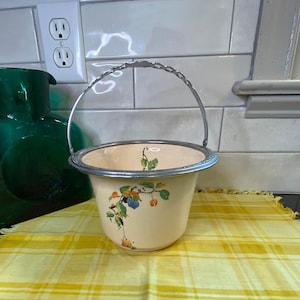 May include: A white ceramic ice bucket with a silver metal handle and floral design. The bucket is sitting on a yellow and white checkered tablecloth.