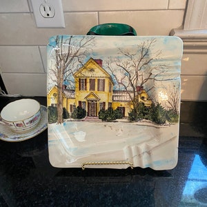 May include: A white ceramic ashtray with a painted image of a yellow house with trees and snow. The ashtray has a gold metal stand.