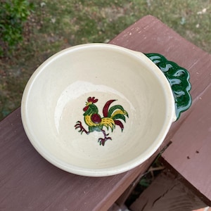 May include: A cream-coloured ceramic bowl with a speckled finish and a hand-painted rooster illustration in the centre. The rooster features red, yellow, and green plumage. The bowl has a dark green, scalloped-edge handle extending from one side.