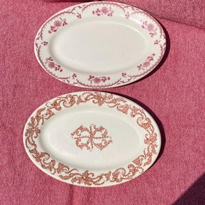 May include: Two oval serving platters with white backgrounds and decorative borders. One platter has a pink floral border, and the other has a brown ornate border. The platters are on a pink surface.