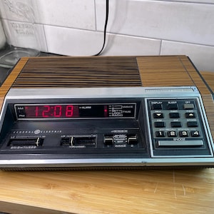 May include: Vintage General Electric programmable clock radio with a wood grain finish. The digital display shows 12:08 in red. Features include alarm, sleep, and radio functions. The clock radio is on a wooden surface.