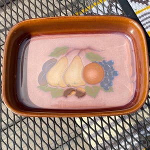 Puede incluir: Plato de cerámica rectangular con borde marrón e interior rosa. El plato presenta un diseño pintado de peras, uvas y una naranja. El plato está sobre una superficie metálica.