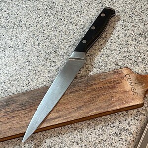 May include: A stainless steel knife with a black handle lies on a wooden sharpening paddle. The paddle is brown with the word "COARSE" stamped on it. A braided leather strap is attached to the paddle.