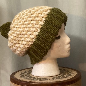 May include: A knitted beanie hat with a white and green colour block design. The hat has a pompom on top.