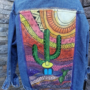 May include: A denim jacket with a hand-painted design on the back. The design features a cactus, a sun, and geometric patterns in shades of orange, yellow, green, purple, and pink.