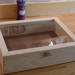 May include: A wooden display case with a glass top and a hinged lid. The case has a silver latch and is made of light wood.